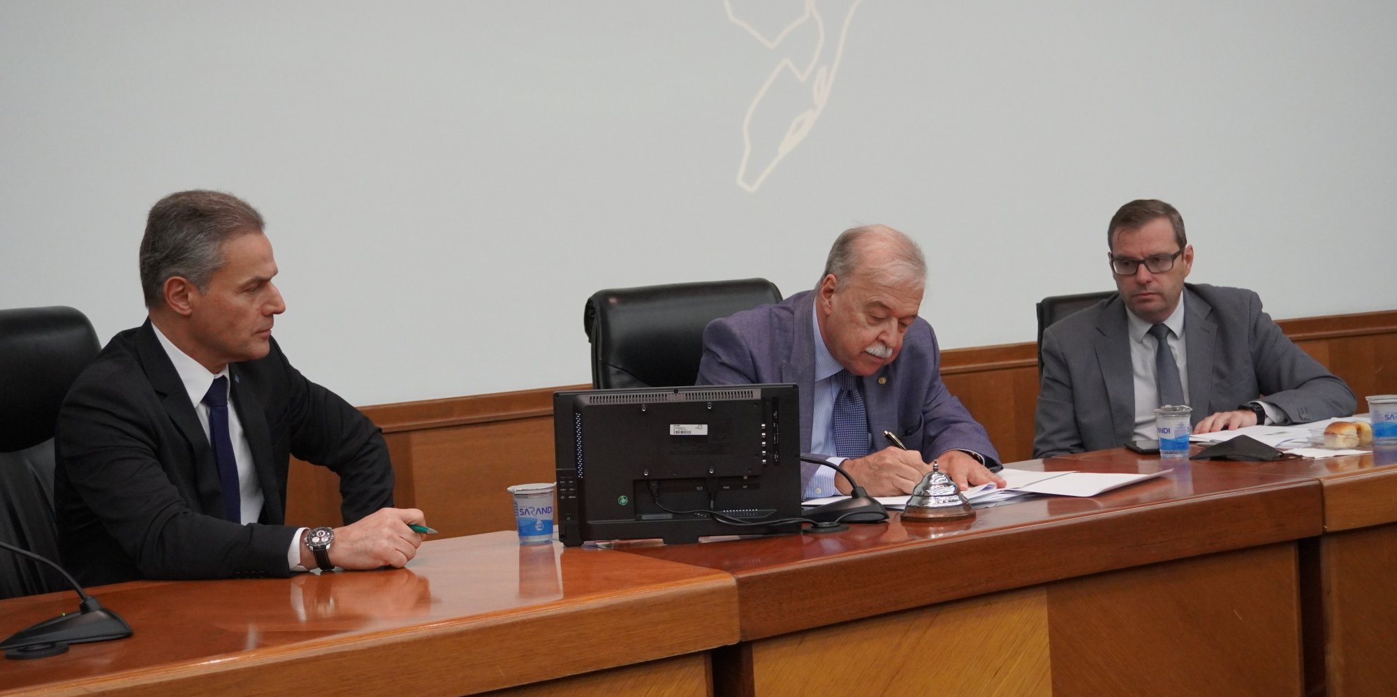 Parede branca, à frente, uma bancada em madeira com três homens brancos sentados, o do meio com uma caneta na mão assinando um documento