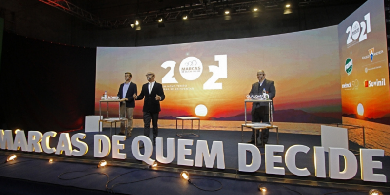 Imagem de um palco, com um letreiro escrito Marcas de Quem Decide 2021 com três homens vestindo terno, um deles em um púlpito