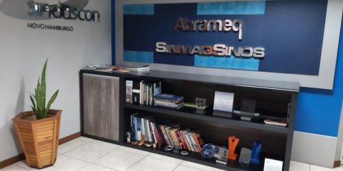 Recepção com parede azul, um quadro escrito Abrameq e SinmaqSinos, uma televisão e uma planta